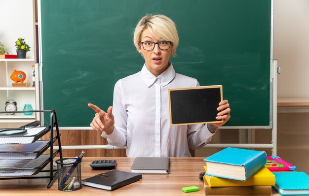 impressed young blonde female teacher wearing glasses sitting at desk with school supplies in classroom showing mini blackboard looking at front pointing at side