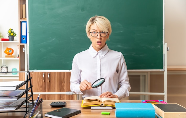 Impressionato giovane insegnante bionda con gli occhiali seduto alla scrivania con gli strumenti della scuola in aula tenendo la lente d'ingrandimento tenendo la mano sul libro aperto