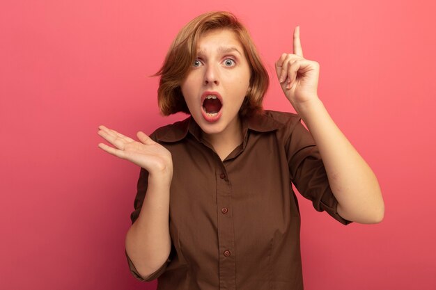 Impressed young blonde girl looking  showing empty hand pointing up isolated on pink wall