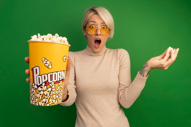 Impressed young blonde girl wearing sunglasses stretching out bucket of popcorn and holding popcorn pieces isolated on green wall