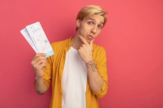 Free photo impressed young blonde guy wearing yellow t-shirt holding tickets and grabbed chin