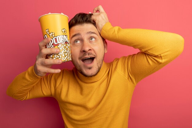 Impressed young blonde handsome man holding bucket of popcorn and popcorn piece touching head with bucket of popcorn and hand looking up isolated on pink wall