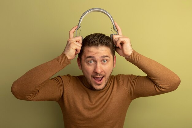 Free photo impressed young blonde handsome man holding headphones above head looking at front isolated on olive green wall