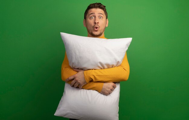 Impressed young blonde handsome man hugging pillow looking straight 