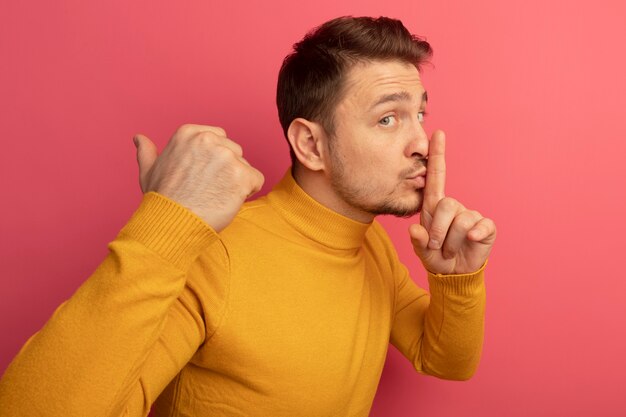 Impressed young blonde handsome man standing in profile view looking at front doing silence gesture pointing behind isolated on pink wall