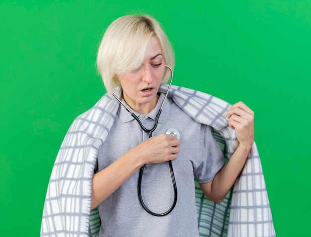 Impressed young blonde ill slavic woman wrapped in plaid holds stethoscope isolated on green wall with copy space