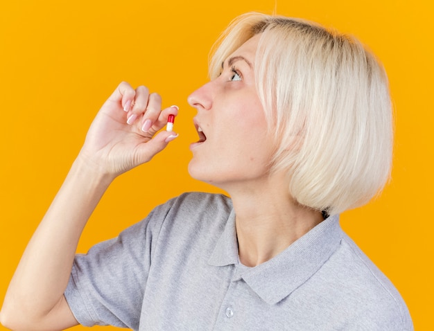 Impressed young blonde ill woman takes medical pill looking at side isolated on orange wall