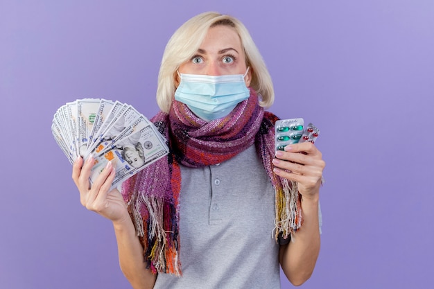 의료 마스크와 스카프를 착용하는 감동 된 젊은 금발의 아픈 여자는 돈과 보라색 벽에 고립 된 의료 약의 팩을 보유
