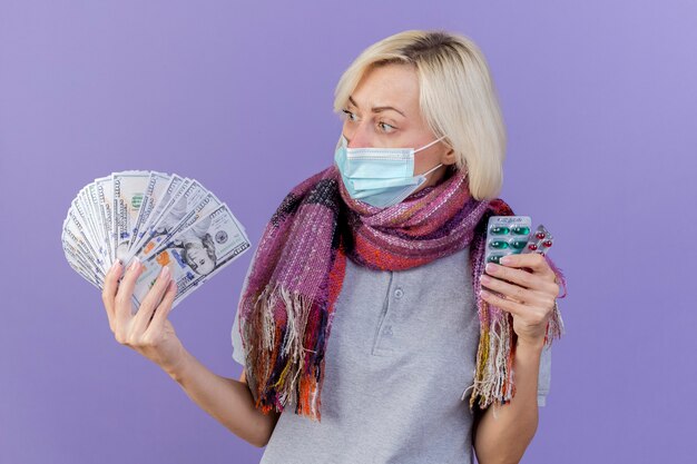 Impressed young blonde ill woman wearing medical mask and scarf holds packs of medical pills looks at money isolated on purple wall