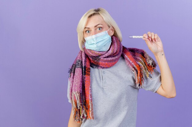 Впечатленная молодая блондинка больная женщина в медицинской маске и шарфе держит термометр, изолированный на фиолетовой стене