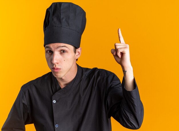 Впечатленный молодой белокурый мужчина-повар в униформе шеф-повара и кепке смотрит в камеру, направленную вверх, изолированную на оранжевой стене