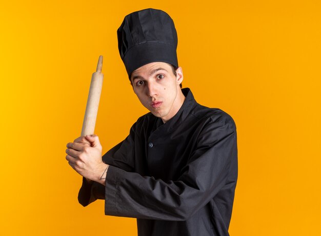 Impressed young blonde male cook in chef uniform and cap standing in profile view holding rolling pin with both hands 