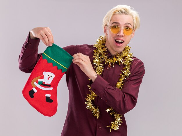 Impressed young blonde man wearing glasses with tinsel garland around neck holding christmas stocking looking at camera isolated on white background