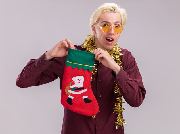 Impressed young blonde man wearing glasses with tinsel garland around neck holding christmas stocking looking at camera isolated on white background