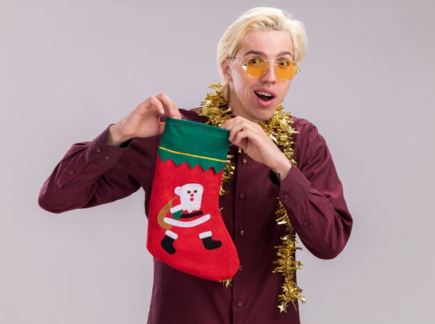 Впечатленный молодой блондин в очках с гирляндой из мишуры на шее, держащий рождественский чулок, глядя в камеру, изолированные на белом фоне