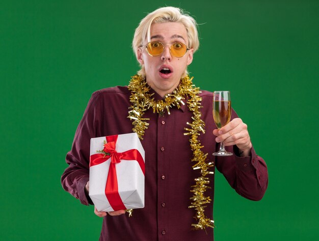 Impressed young blonde man wearing glasses with tinsel garland around neck holding glass of champagne and gift package  isolated on green wall