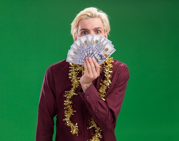 Впечатленный молодой блондин в очках с гирляндой из мишуры на шее, держащий деньги сзади, изолированные на зеленой стене с копией пространства