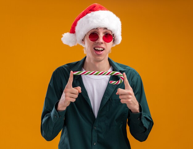 오렌지 배경에 고립 된 카메라를 가로로보고 크리스마스 달콤한 지팡이를 들고 산타 모자와 안경을 쓰고 감동 젊은 금발의 남자