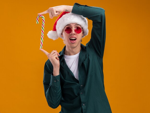 コピースペースでオレンジ色の背景に分離されたカメラを垂直に見てクリスマスの甘い杖を保持しているサンタの帽子と眼鏡を身に着けている印象的な若いブロンドの男