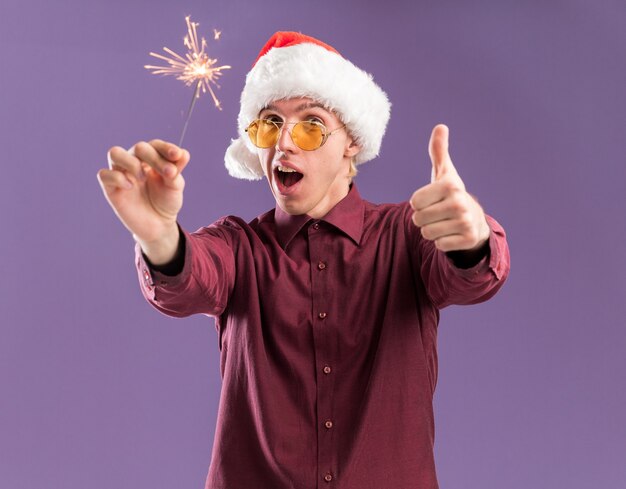 Impressed young blonde man wearing santa hat and glasses holding holiday sparkler  showing thumb up isolated on purple wall