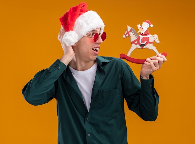 Impressionato giovane uomo biondo che indossa cappello e occhiali da babbo natale tenendo e guardando santa sulla figurina di cavallo a dondolo tenendo la mano dietro la testa isolata su sfondo arancione