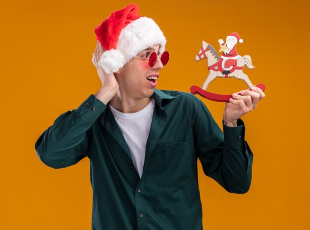 Impressed young blonde man wearing santa hat and glasses holding and looking at santa on rocking horse figurine keeping hand behind head isolated on orange background