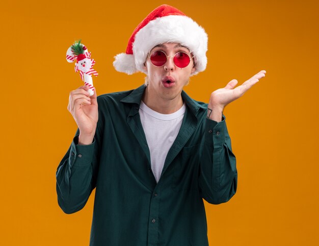 오렌지 배경에 고립 된 빈 손을 보여주는 카메라를보고 달콤한 지팡이 장식을 들고 산타 모자와 안경을 착용하는 감동 된 젊은 금발의 남자