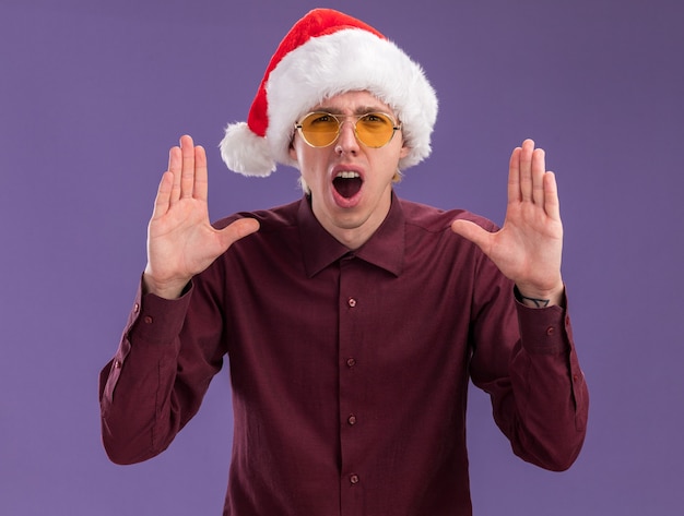 Impressionato giovane uomo biondo che indossa cappello santa e occhiali guardando la telecamera tenendo le mani in aria gridando ad alta voce qualcosa di isolato su sfondo viola