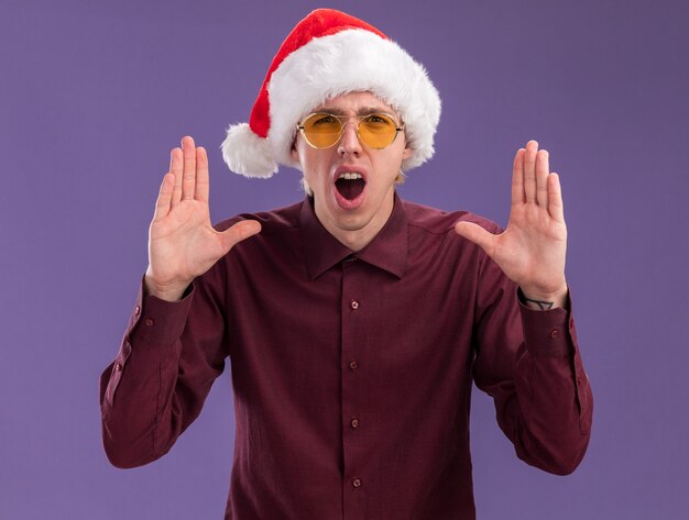 Impressed young blonde man wearing santa hat and glasses looking at camera keeping hands in air shouting out loud something isolated on purple background