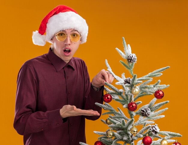 오렌지 배경에 고립 된 카메라를보고 트리를 가리키는 크리스마스 지팡이를 들고 장식 된 크리스마스 트리 근처에 서있는 산타 모자와 안경을 착용하는 감동 된 젊은 금발의 남자