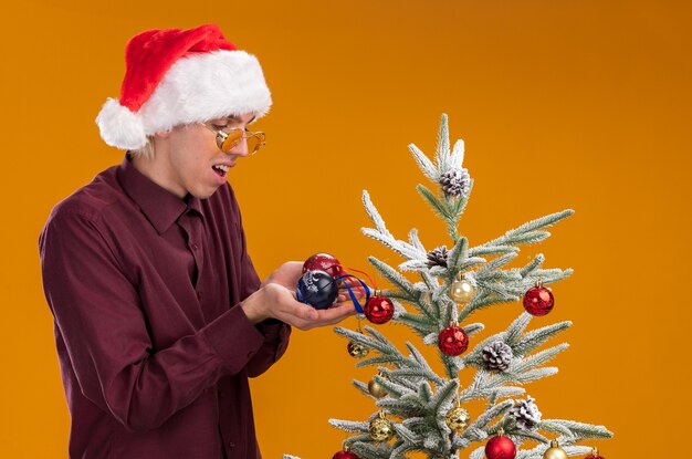 오렌지 배경에 장식 된 크리스마스 트리 근처 프로필보기에 서 산타 모자와 안경을 착용하는 감동 된 젊은 금발의 남자
