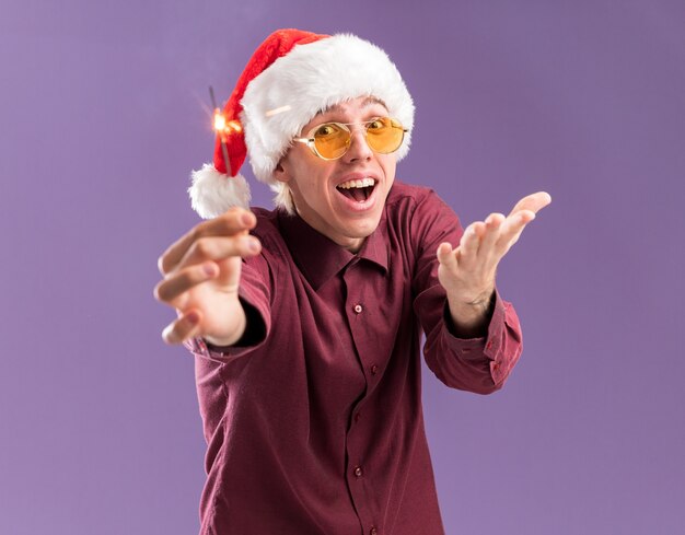 Impressed young blonde man wearing santa hat and glasses stretching out holiday sparkler   showing empty hand isolated on purple wall