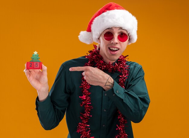 Impressed young blonde man wearing santa hat and glasses with tinsel garland around neck holding and pointing at christmas tree toy with date looking at camera isolated on orange background