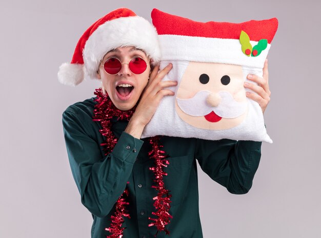 Impressed young blonde man wearing santa hat and glasses with tinsel garland around neck holding santa claus pillow touching head with it looking at camera isolated on white background