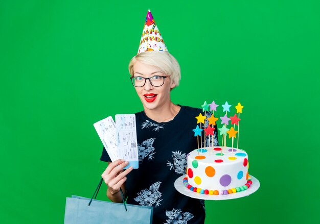 Impressed young blonde party girl wearing glasses and birthday cap holding birthday cake with stars airplane tickets and paper bag looking at camera isolated on green background with copy space