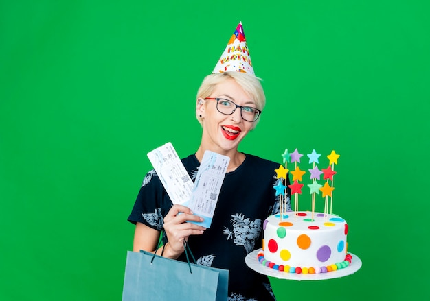 Free photo impressed young blonde party girl wearing glasses and birthday cap holding birthday cake with stars airplane tickets and paper bag looking at camera isolated on green background with copy space