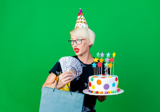 Impressed young blonde party girl wearing glasses and birthday cap holding birthday cake with stars money gift box and paper bag looking at camera isolated on green background with copy space