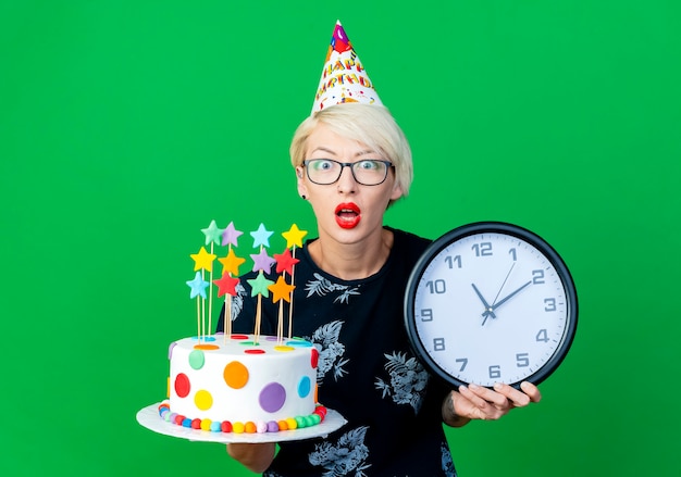 Impressed young blonde party girl wearing glasses and birthday cap holding birthday cap and clock looking at camera isolated on green background with copy space