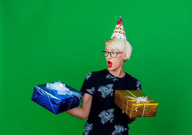 Impressed young blonde party girl wearing glasses and birthday cap holding gift boxes looking at one of them isolated on green background