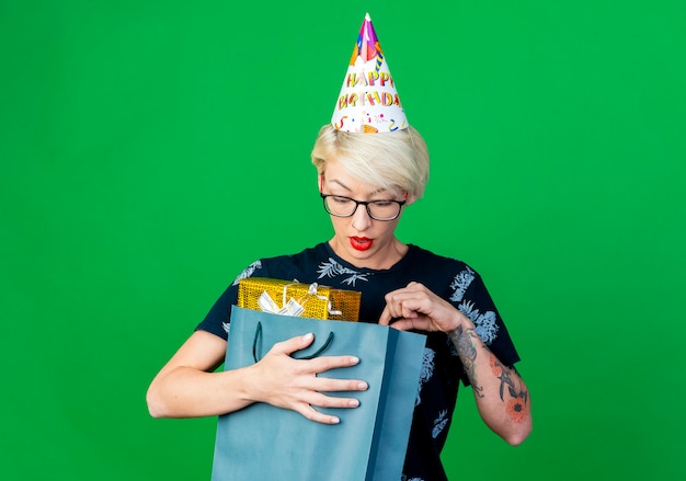 Free photo impressed young blonde party girl wearing glasses and birthday cap holding and looking inside paper bag with gift boxes isolated on green background with copy space