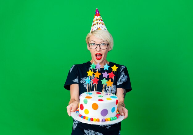Впечатленная молодая белокурая тусовщица в очках и кепке на день рождения, протягивающая торт ко дню рождения со звездами, смотрящая в камеру, изолированную на зеленом фоне с копией пространства