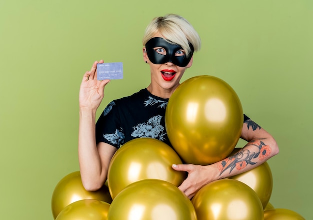 Impressed young blonde party girl wearing masquerade mask standing behind balloons showing credit card looking at camera isolated on olive green background