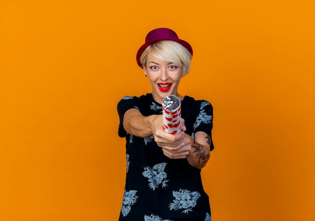 Impressed young blonde party girl wearing party hat looking at camera stretching out confetti cannon towards camera isolated on orange background with copy space