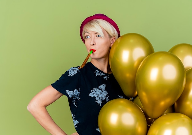 Free photo impressed young blonde party girl wearing party hat standing behind balloons keeping hand on waist looking at side blowing party blower isolated on olive green background