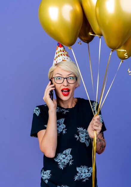 Impressed young blonde party woman wearing glasses and birthday cap holding balloons talking on phone looking at front isolated on purple wall