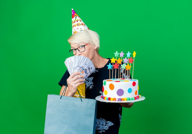 Impressed young blonde party woman wearing glasses and birthday cap holding cake with stars money gift box and paper bag