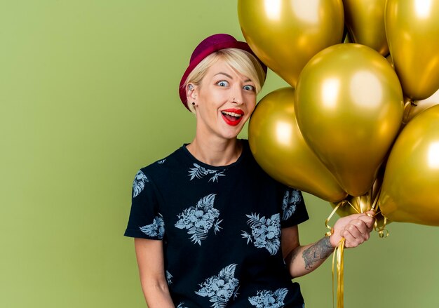 Impressed young blonde party woman wearing party hat holding balloons looking at front isolated on olive green wall with copy space