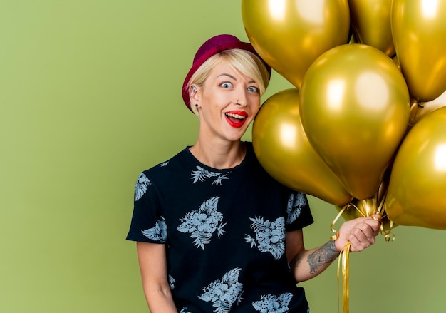 Impressed young blonde party woman wearing party hat holding balloons looking at front isolated on olive green wall with copy space