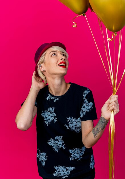 Impressed young blonde party woman wearing party hat holding and looking at balloons touching hat isolated on pink wall