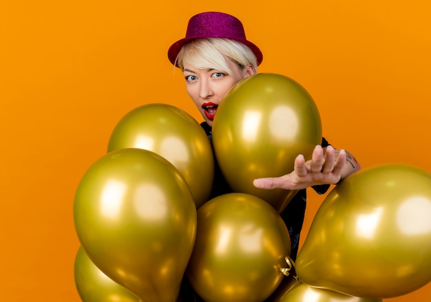 Impressed young blonde party woman wearing party hat standing behind balloons looking at front stretching out hand towards front isolated on orange wall